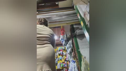 Video of a woman with a medium build and short brown hair, wearing a beige shirt, shopping in a cluttered convenience store. Shelves are lined with various products, including snacks and household items.