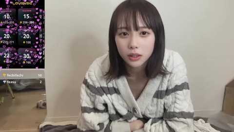 Video of an East Asian woman with shoulder-length black hair and fair skin, wearing a white and gray striped bathrobe, kneeling on a tiled floor. Background includes a calendar and a blurred room.