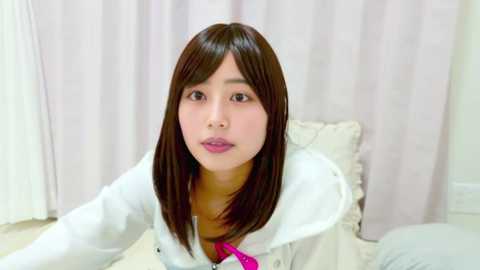 Video of an East Asian woman with straight, shoulder-length brown hair, wearing a white jacket with a pink scarf. She sits on a bed with white pillows against a light pink curtain background.