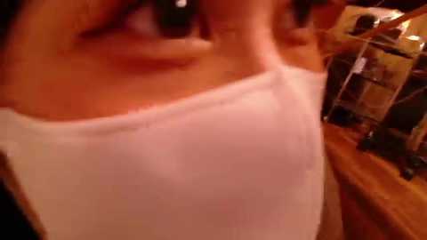 Close-up video of a person wearing a white tank top, with a blurred background featuring wooden shelves and dim lighting, possibly in a home setting.