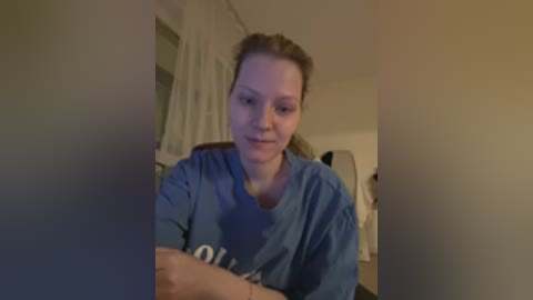 Video of a Caucasian woman with light skin and brown hair tied back, wearing a blue hospital gown, sitting on a bed in a dimly lit room.