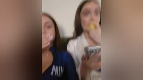 Media: A blurry video captures two young women, one with long brown hair in a dark blue jacket, the other with straight brown hair in a white shirt, eating popcorn, set against a plain background.