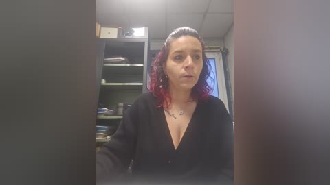 Video of a middle-aged woman with curly red hair, wearing a black cardigan, standing in a cluttered office with bookshelves and a window in the background.