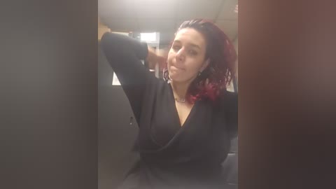 A video of a young woman with medium-length, wavy, red hair, wearing a black dress and black earrings, posing in a dimly lit room with a blurred background.