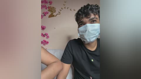Video of a young man with curly hair, wearing a mask and black T-shirt, seated indoors with pink floral wall decor and birds on beige walls.