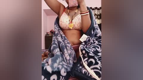 Video of a light-skinned woman with medium-sized breasts, wearing a floral-patterned bra, draped in a blue and white patterned blanket, standing in a cluttered room with a pink wall and dark furniture.