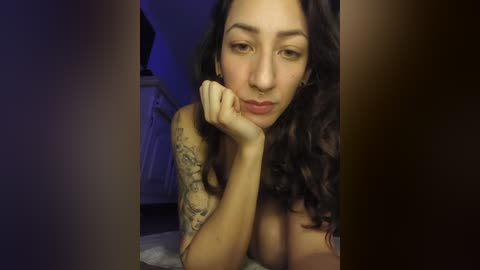 A video of a young, topless woman with dark curly hair, lying on her side, resting her chin on her hand. She has a tattoo on her arm and a nose ring.