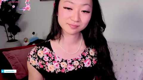 A video of an Asian woman with long black hair and fair skin, wearing a floral dress, sitting in a bedroom with pink bedding and a lamp.