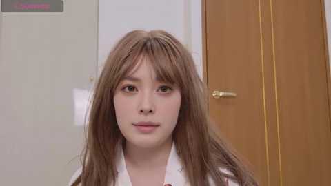 Video of a young East Asian woman with long, straight brown hair, wearing a white shirt, standing indoors near a wooden door.