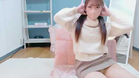 A video of an Asian woman with straight, brown hair in pigtails, wearing a white turtleneck sweater and a short, brown skirt, sitting on a plush, pink chair in a room with light blue walls and wooden floors.