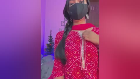 Video of a woman with dark skin, wearing a red and white patterned kurta, black face mask, and long black braid, standing indoors with purple and red lighting.