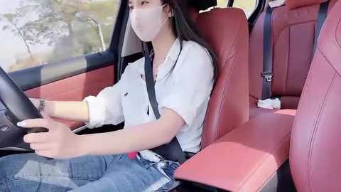 Video of an Asian woman in a white blouse and blue jeans driving a red car, wearing a face mask.