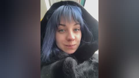 Video of a young woman with pale skin, shoulder-length blue hair, and green eyes, wearing a black faux fur hood, smiling. Background is blurred.