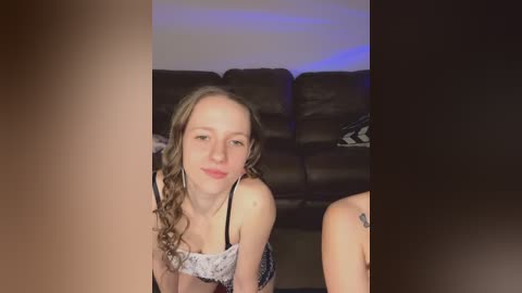 Video of a young Caucasian woman with wavy brown hair, fair skin, and a slender physique, wearing a black lace lingerie top, smiling and kneeling on a dark sofa with a patterned cushion in a dimly lit room.