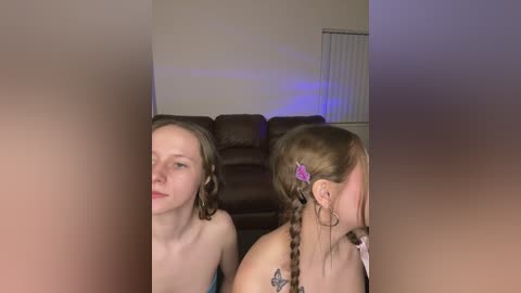 Video of two young women with light skin, one topless, seated on a brown leather couch. One has braided hair adorned with a purple hair clip, the other with loose hair.