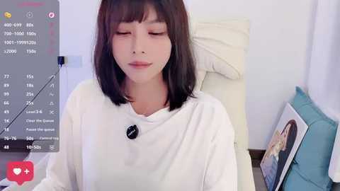 Video of a young East Asian woman with straight, shoulder-length dark brown hair, wearing a white turtleneck sweater, eyes closed, in a cozy bedroom with a bed and teal pillow.
