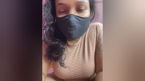 Video of a woman with medium brown skin, wearing a black face mask, a light beige t-shirt, and a pink background.