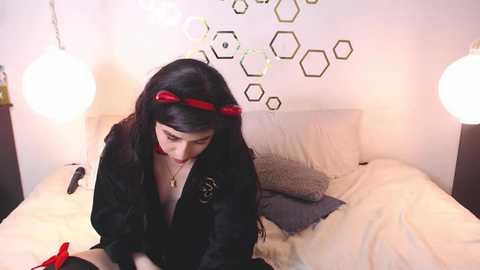 Video of a young woman with long black hair, wearing a black robe with red accents, sitting on a bed in a modern, hexagonal-patterned room with white walls and two spherical lamps.