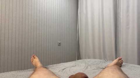 A video showing a person's bare feet and lower legs lying on a light-colored bed with a patterned sheet, against a wall with vertical striped wallpaper and a white curtain.