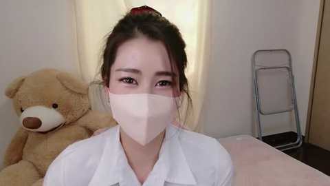 Video of an Asian woman with light skin and dark hair tied up, wearing a white medical mask, standing indoors near a beige curtain and a wooden chair with a teddy bear.
