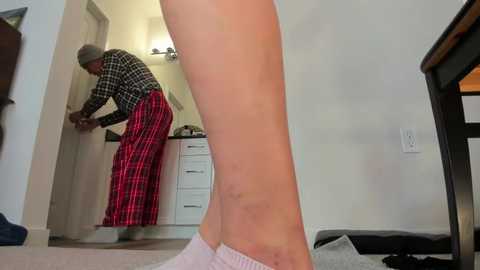 Video of an elderly Black woman in a plaid skirt, bending over, in a cozy, well-lit living room with a beige carpet and white walls.