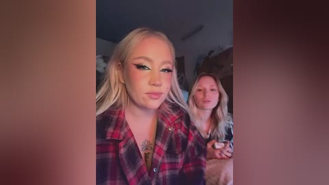 Video of two young women, one blonde with a red plaid shirt, the other with long hair and a gray sweater, smiling in a dimly lit room with brown vertical lines.