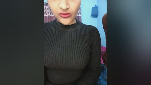 Video of a woman with light skin, wearing a tight black ribbed turtleneck, showing moderate cleavage, and red lipstick. Background features a pink and white patterned curtain and a blue wall with a hook.