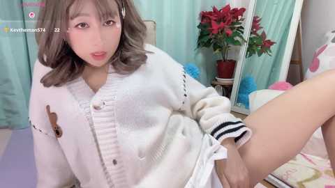 A video of an Asian woman with shoulder-length, wavy, gray hair, wearing a white cardigan and white shorts, posing seductively on a bed with potted plants and pastel-colored curtains in the background.