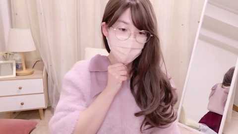 Video of an East Asian woman with long, wavy brown hair, wearing glasses and a lavender bathrobe, holding her chin, in a softly lit, minimalist bedroom with white walls, a mirror, and a nightstand.