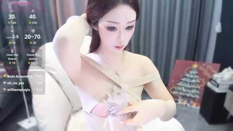 Video of an East Asian woman with fair skin and long brown hair, wearing a beige coat and holding a glass. Background includes a Christmas tree and a social media overlay.