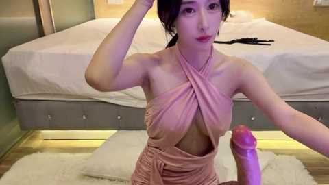 Video of an Asian woman with fair skin, wearing a revealing, pink halter top, sitting on a white rug in a modern bedroom with a gray upholstered bed and wooden floors.