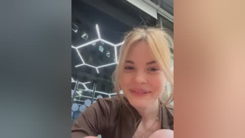 Video of a smiling, fair-skinned, blonde woman with light makeup, wearing a brown jacket, standing in front of a hexagonal light installation in a modern, industrial-style room.
