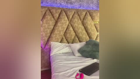 Video of a cozy bedroom with a tufted, diamond-patterned gold headboard, a white duvet, teal pillow, and a laptop on the bed.