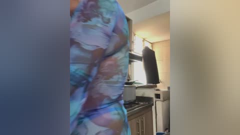 Video of a person wearing a colorful, abstract-patterned dress in a modern kitchen with light-colored cabinets, a stove, and a black jacket hanging on the wall.