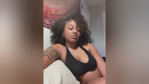 Video of a young Black woman with a large afro, lying on a bed with a black sports bra, visible tattoos on her left arm, and a tapestry in the background.