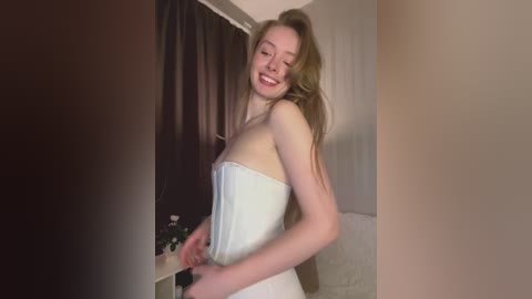 A video of a smiling, fair-skinned, slender woman with long, light brown hair, wearing a white strapless dress, standing in a dimly lit room with beige walls and a white couch.