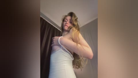 Video of a light-skinned woman with long, wavy blonde hair, wearing a white tank top, lifting it to expose her small breasts. She stands in a dimly lit room with brown curtains and beige walls.