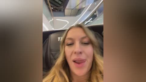 A video captures a smiling, light-skinned woman with long, wavy blonde hair, seated in a black leather chair, possibly inside a vehicle or airplane, with a blurred, colorful background.