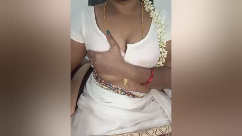 Video of a South Asian woman with medium-dark skin, wearing a white cropped blouse, floral-patterned skirt, and a garland of white flowers. She has a gold necklace and red bracelets. Her left hand covers her breast.