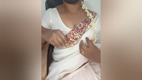 Video of a person with medium brown skin wearing a white saree adorned with a colorful floral border, adjusting a blue face mask. The background is blurred.