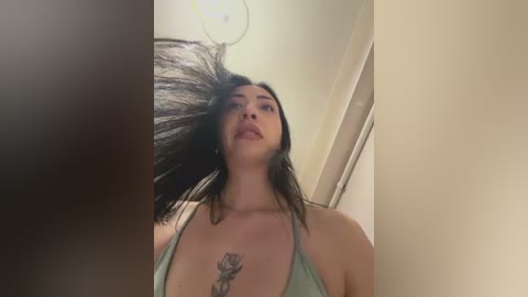 Video of a woman with long black hair, wearing a green tank top, standing in a beige room with a white door and a ceiling light.