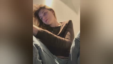 A video of a young woman with long brown hair, lying on a bed, wearing a brown top and light blue jeans. The image has a soft, warm lighting and a blurred background.