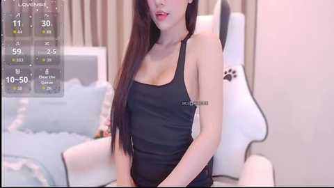 A video of an Asian woman with long black hair, wearing a black tank top, sitting on a white gaming chair in a softly lit bedroom.