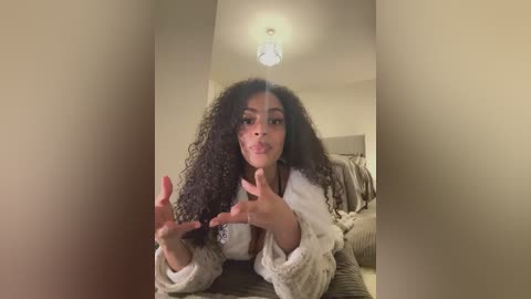 A video of a young Black woman with voluminous curly hair, wearing a white robe, making a heart gesture with her hands in a dimly lit room with beige walls and a chandelier.