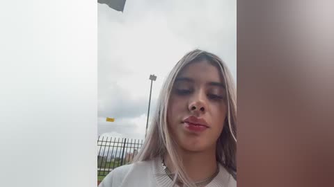 Video of a young woman with long, straight, light brown hair, wearing a white sweater and a gold necklace, standing outdoors near a metal fence. Background shows a cloudy sky and a yellow light pole.