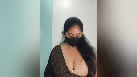Video of a South Asian woman with long black hair, wearing a black mask, brown knit top, and beige bra, standing in a minimalistic room with light blue and white walls.