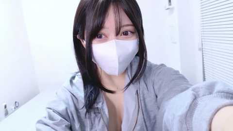 A video of a young East Asian woman with straight black hair, wearing a white face mask, light gray hoodie, and revealing a hint of cleavage, taken indoors in a sterile, white room with blinds.