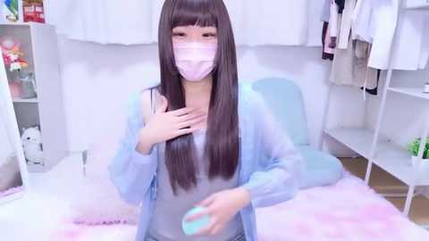Video of a young Asian woman with long brown hair and pale skin, wearing a pastel blue robe, mask, and light blue nail polish, sitting on a fluffy bed in a soft-lit, pastel room.