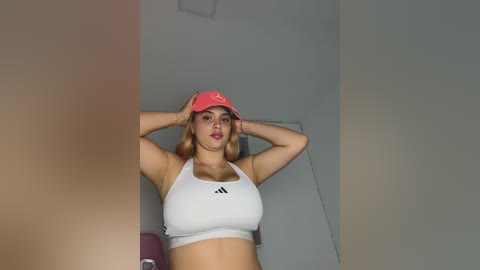 A video of a young woman with fair skin, wearing a white sports bra and pink baseball cap, standing against a plain white wall.