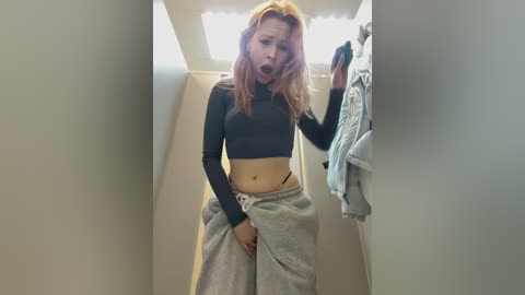 A young woman with long, wavy red hair, wearing a black crop top and grey sweatpants, is taking a selfie in a narrow hallway. She has a surprised expression and her hand is on her crotch.